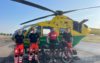 A group of people standing in front of an air ambulance helicopter. Three of the are clinical crew and one is a cyclist, who is standing with his bicycle.