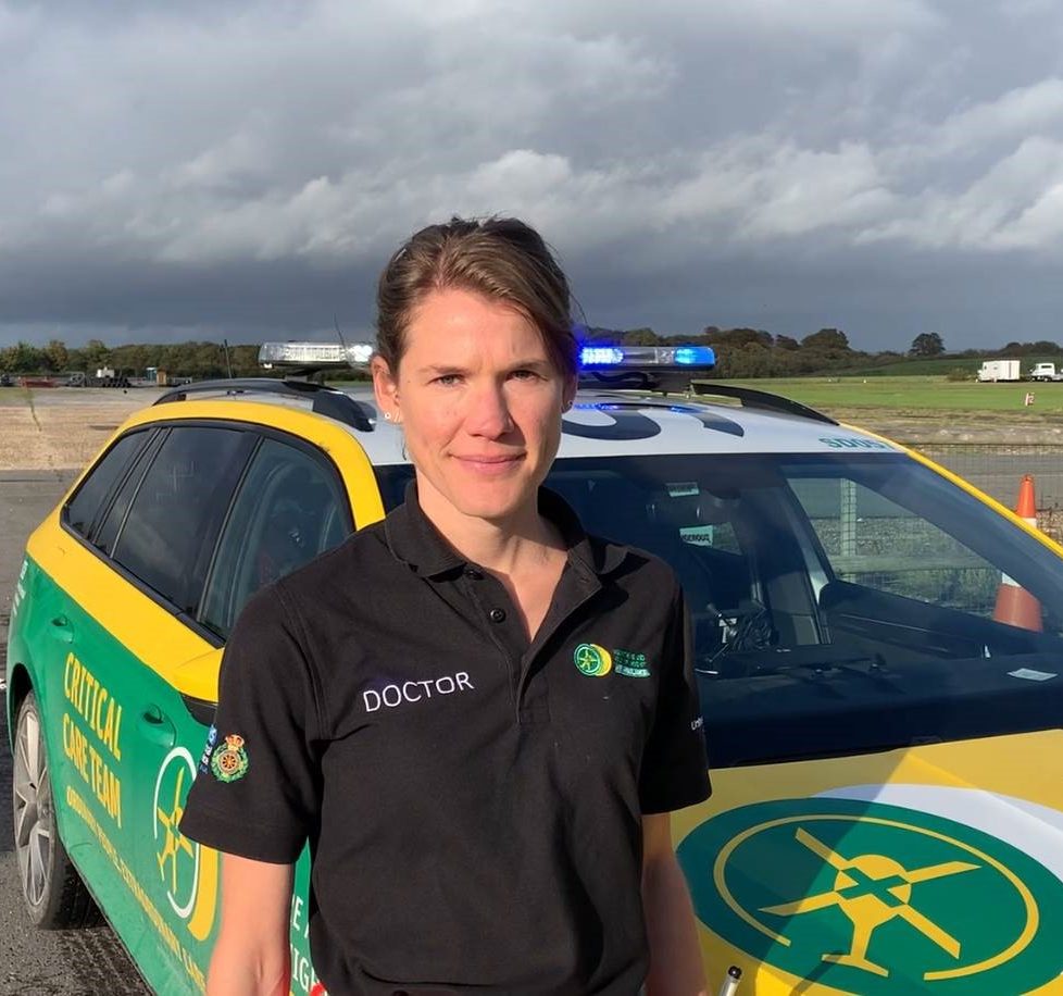 Dr Frances Dolman is wearing a black polo Doctor polo shirt with the Hampshire and Isle of Wight Air Ambulance logo. She is stood in front of a HIOWAA emergency response vehicle.