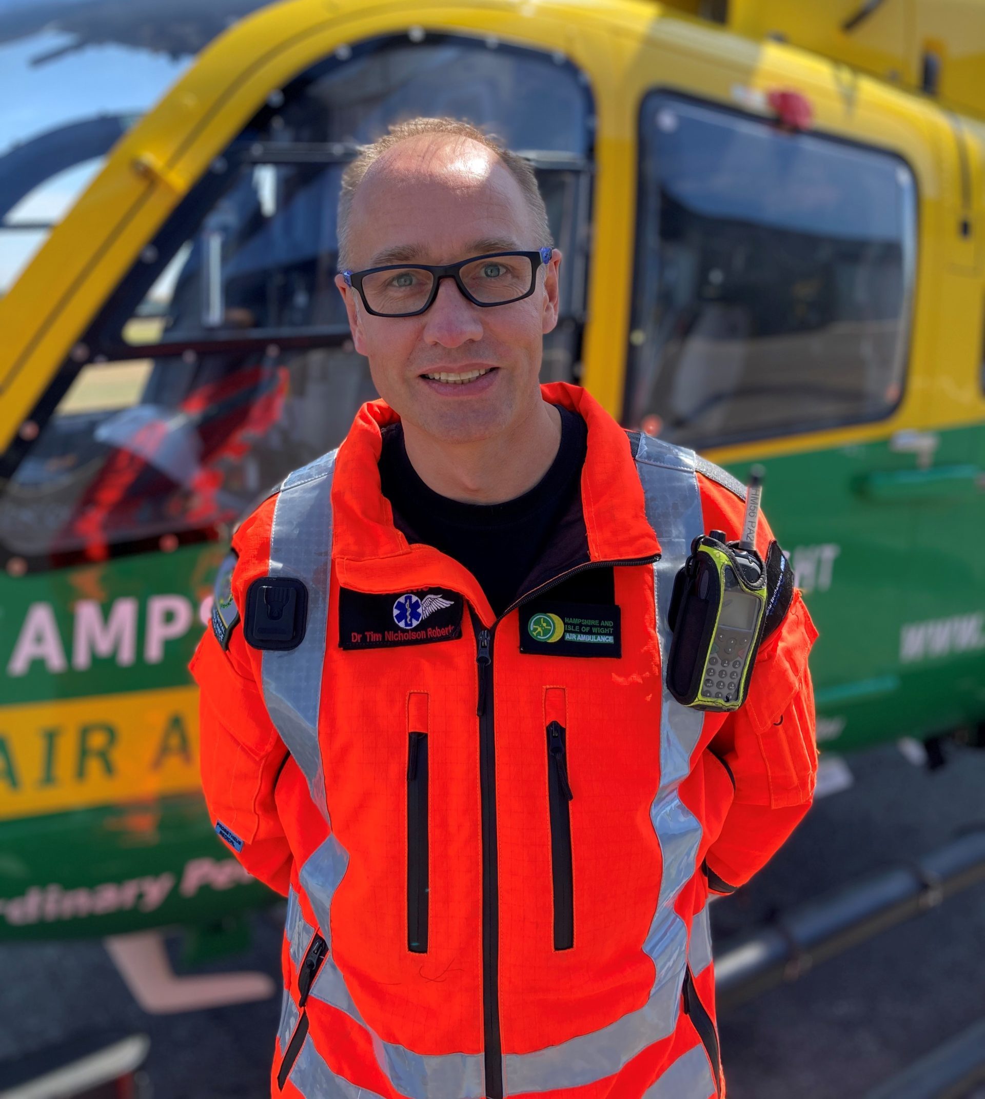 Dr Tim Nicholson Roberts is smiling to the camera. The helicopter is behind him.