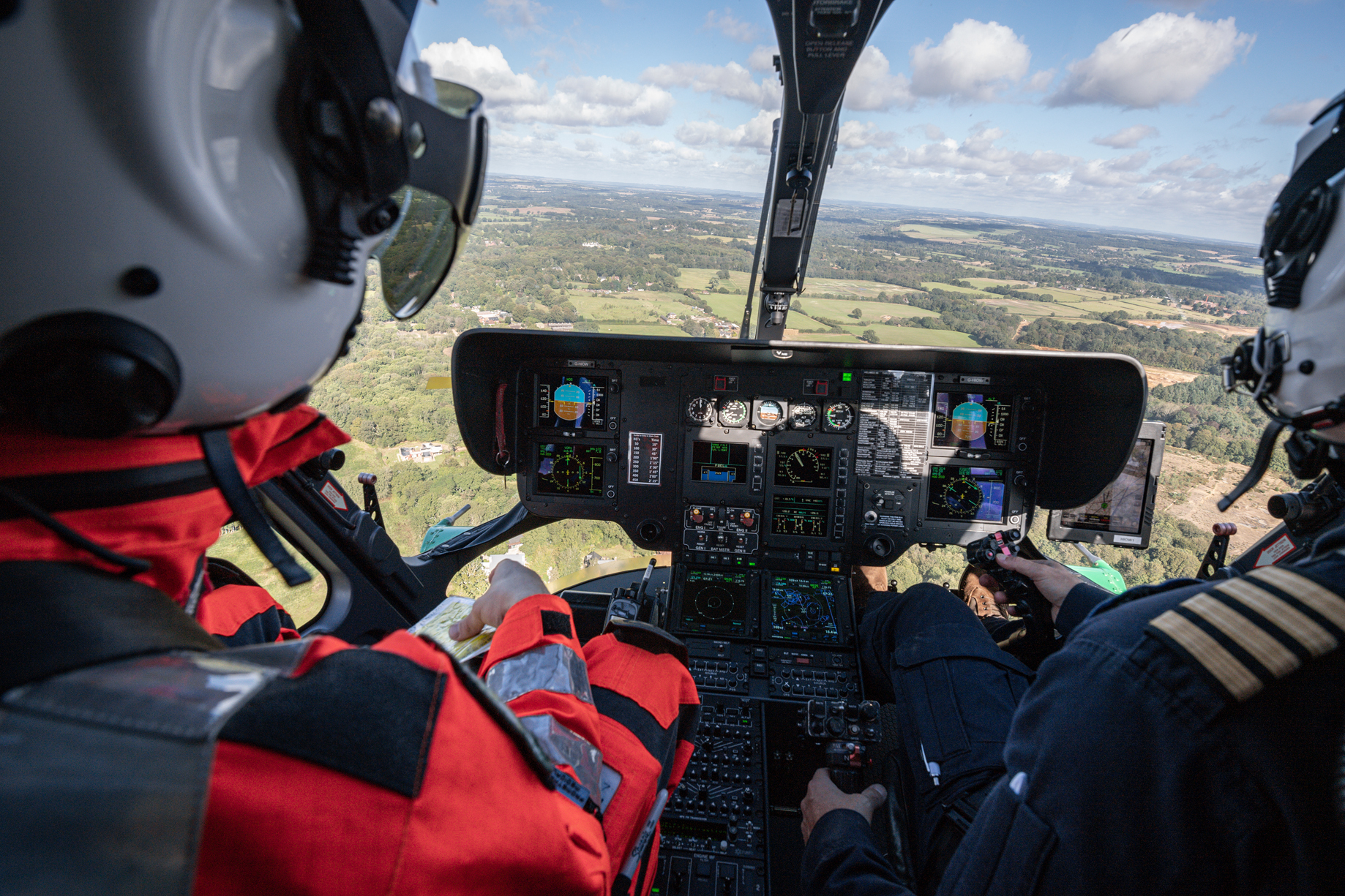 View from the inside of the helicopter looking out of the front. There is a paramedic to the left and the pilot to the right. We are flying over fields.