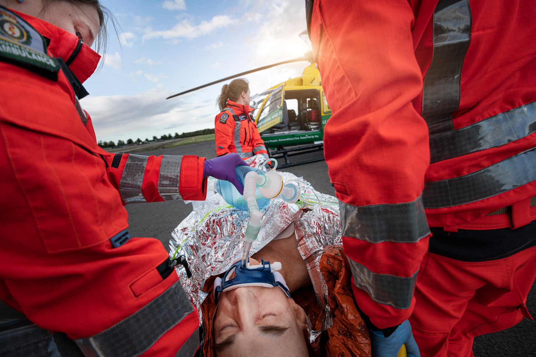 Three crew members are participating in a training simulation with a life like manikin. The manikin is on already packed onto a stretcher and is wearing an oxygen mask. The helicopter is in the background.