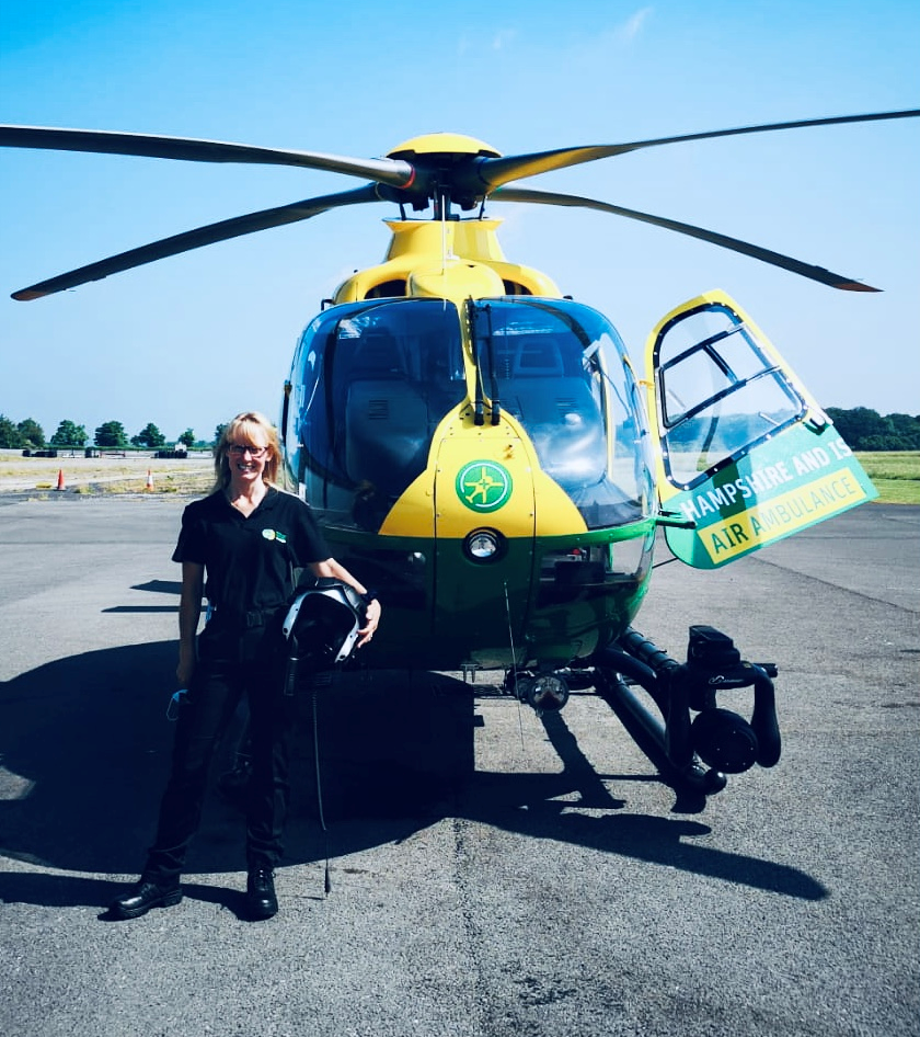 A lady holding a flight helmet stood in front of a yellow and green helicopter with one of its doors open