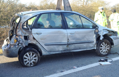 The side view of a silver car that has both the back and front end smashed and the rear tyre blown out