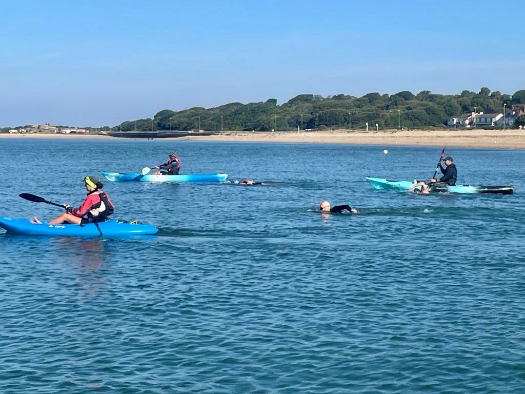 A group of kayakers and swimmers in the water with a beach in the background