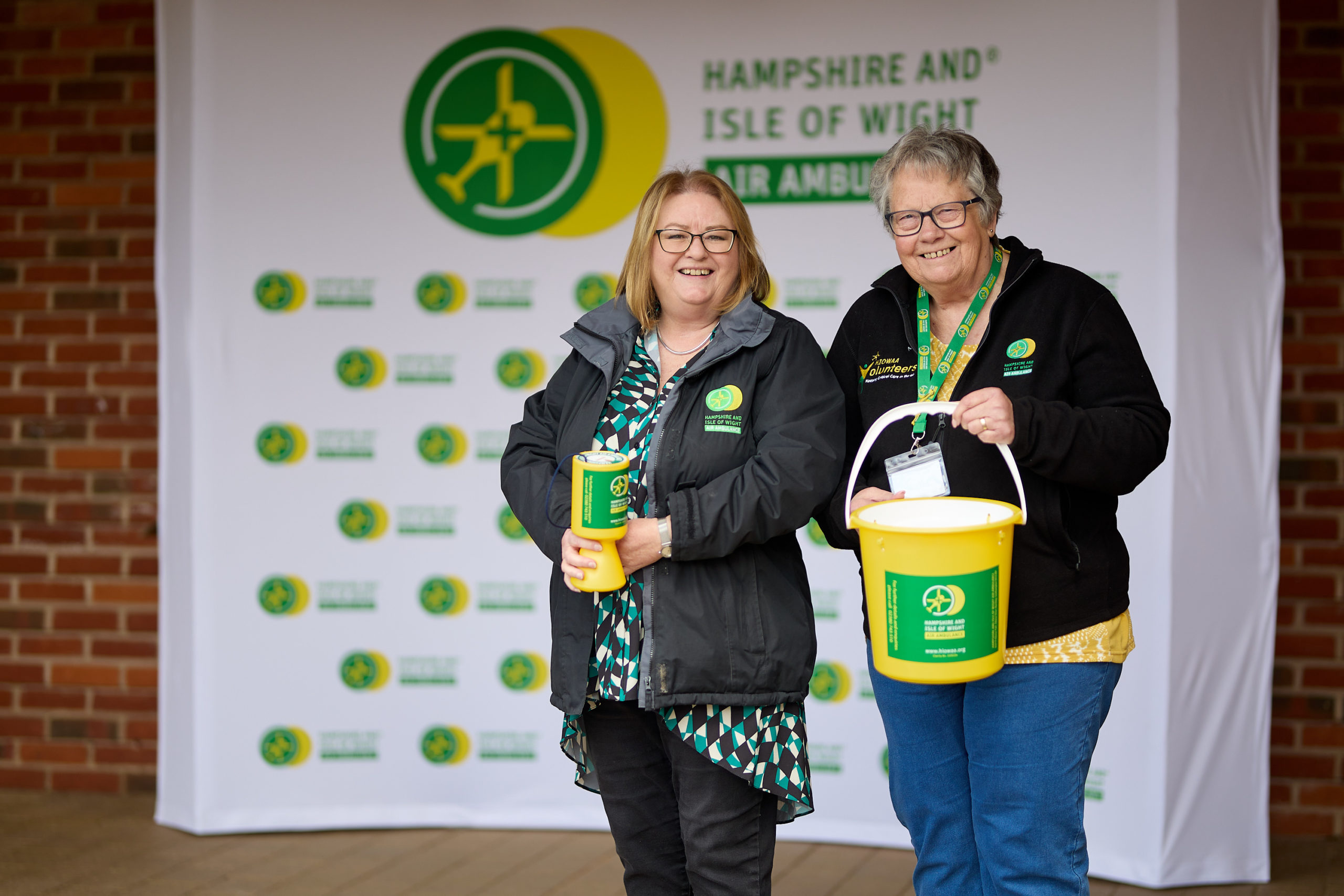 Two volunteers are preparing to assist at an event for the charity. They are carrying a collection pot and bucket and are both smiling