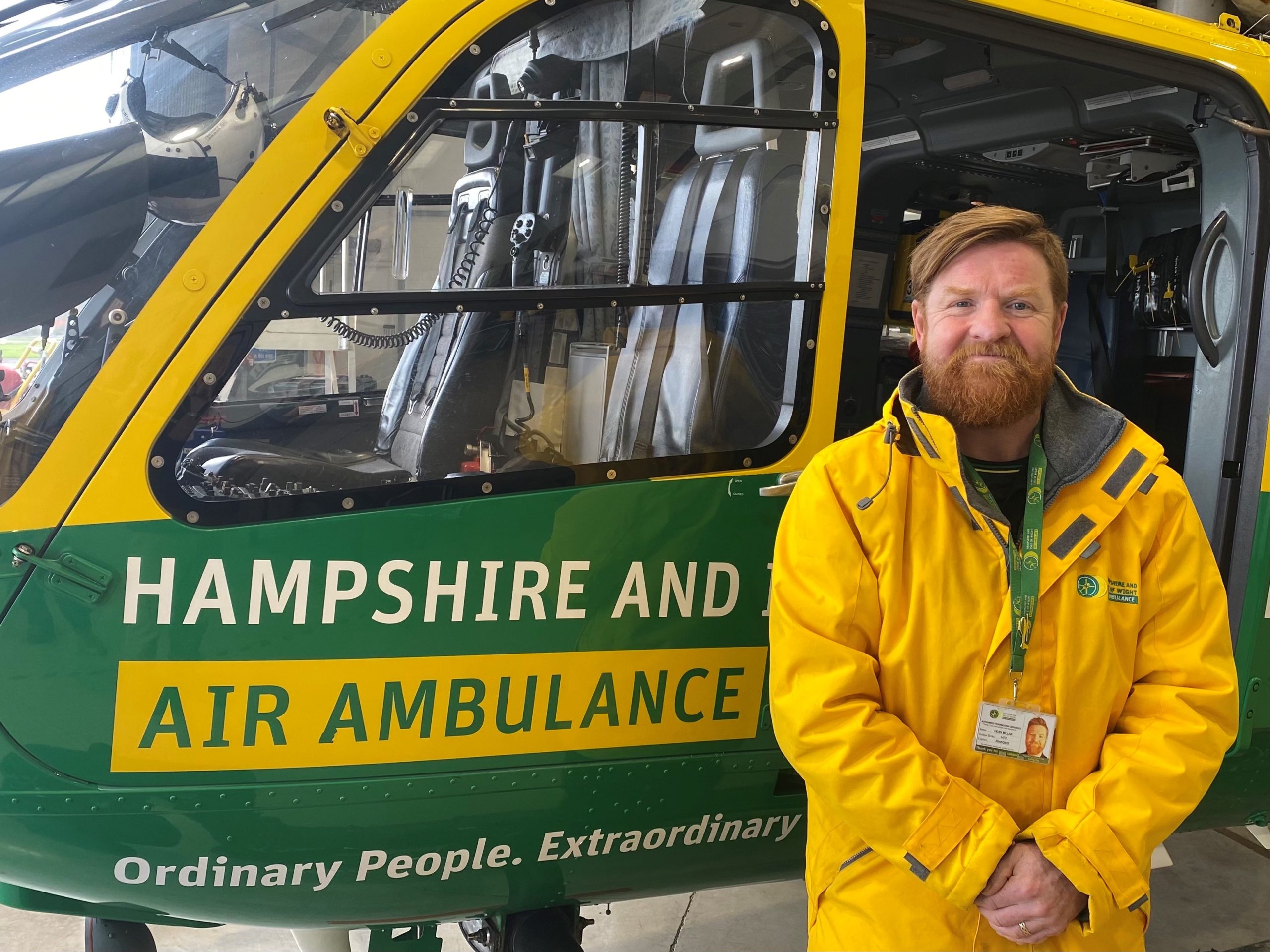 Dean is standing in front of the helicopter in the hangar. He is wearing a yellow rain jacket and is smiling to camera.