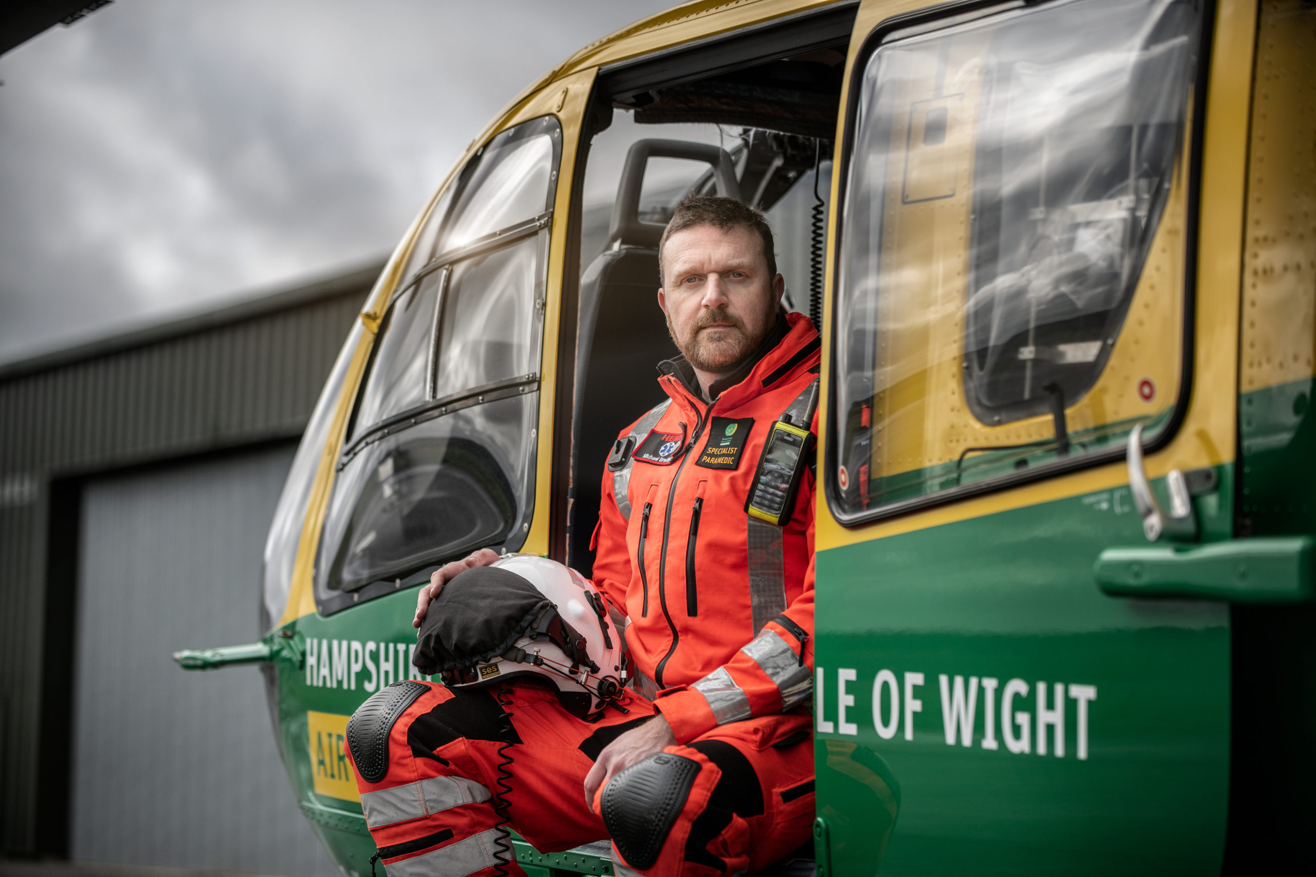 HEMS Paramedic Lead, Michael Bradfield, is sat in the Helicopter in his orange flight suit, looking at the camera. His flight helmet is resting on his leg.