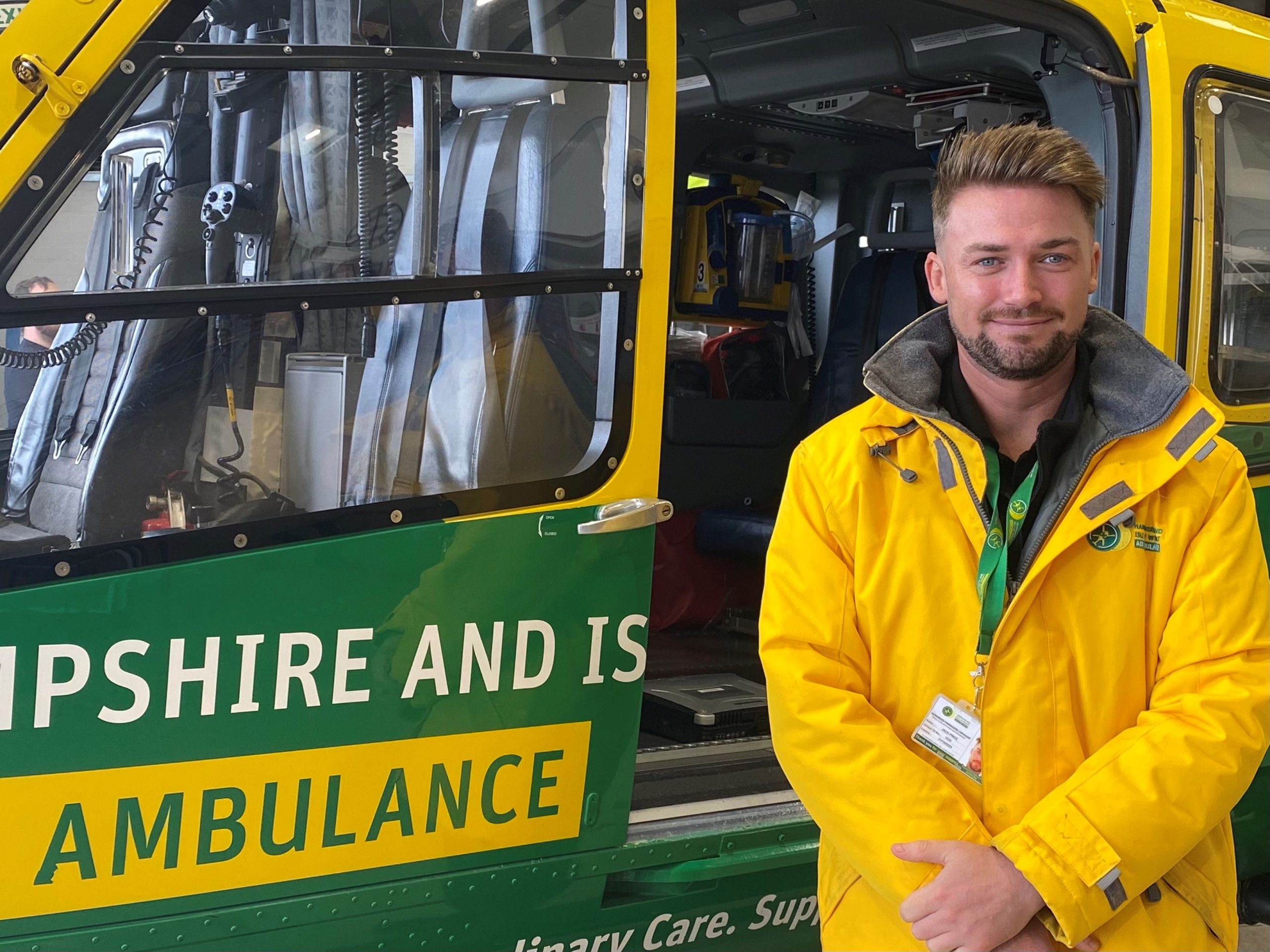 Jack is standing in front of the helicopter in the hangar. He is wearing a yellow rain jacket and is smiling to camera.