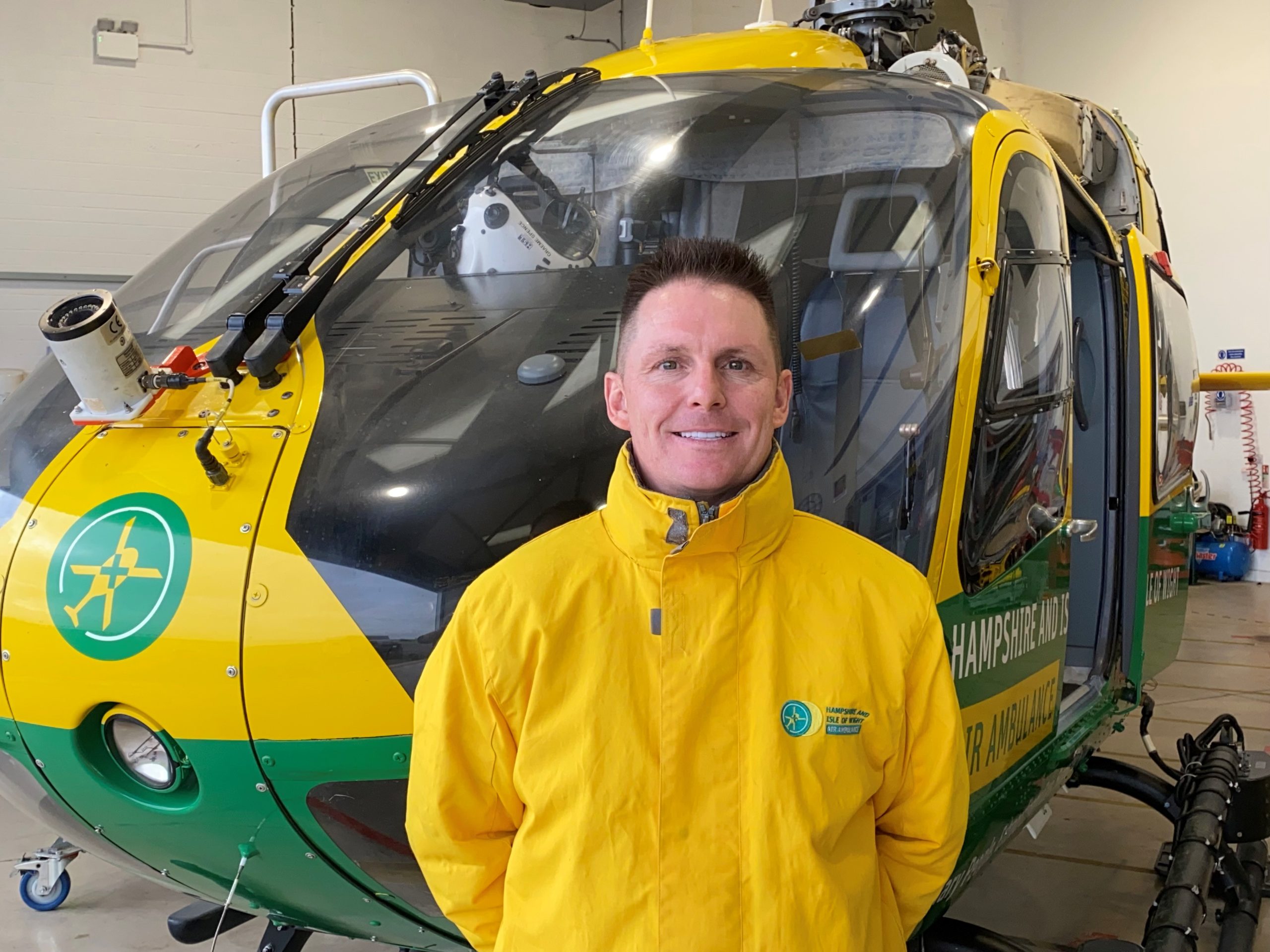 Seb is standing in front of the helicopter in the hangar. He is wearing a yellow rain jacket and is smiling to camera.