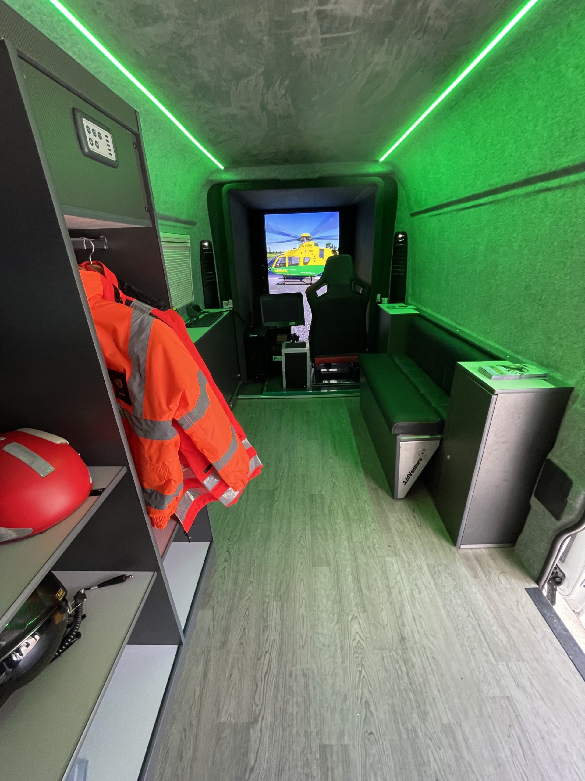 Inside a van that has been converted into a flight simulator. Orange aircrew flight jackets and helmets are hanging up.