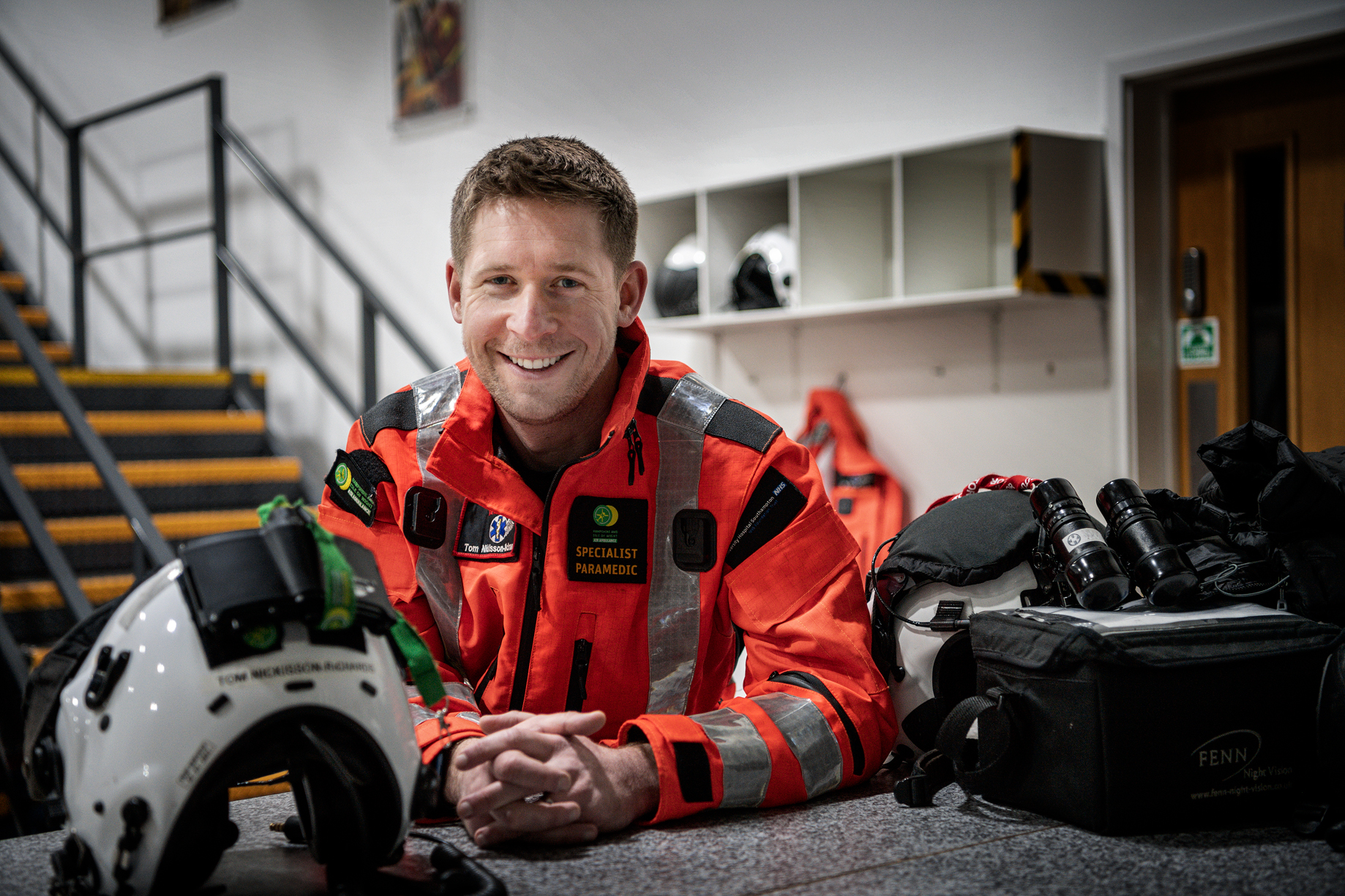 Specialist Paramedic Tom is in his flight suit in the hanger, smiling at to the camera. His flight helmet is to the side.