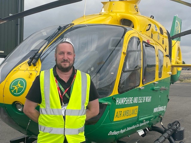 Russell is standing in front of the green and yellow helicopter. He is smiling to camera and is wearing his uniform and a high visibility jacket.