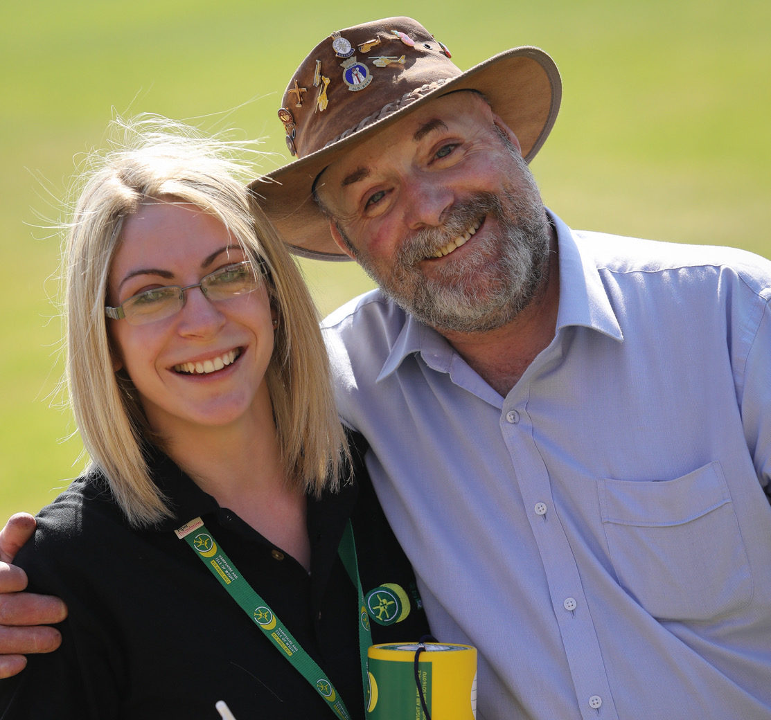 A man and woman looking at the camera smiling. They're holding a charity collection pot.