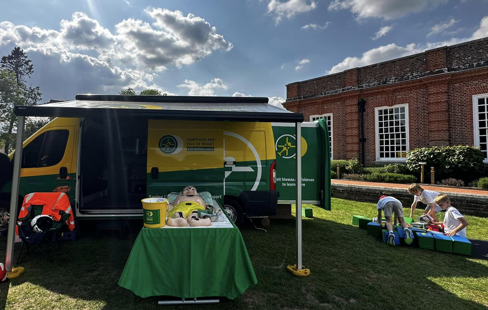The Hampshire and Isle of Wight Simulation Van is set up at an event. The side door is open and a giant Operation game is on a table at the front, with a donation bucket on top. Three children are playing with the cube puzzle to the right hand side.