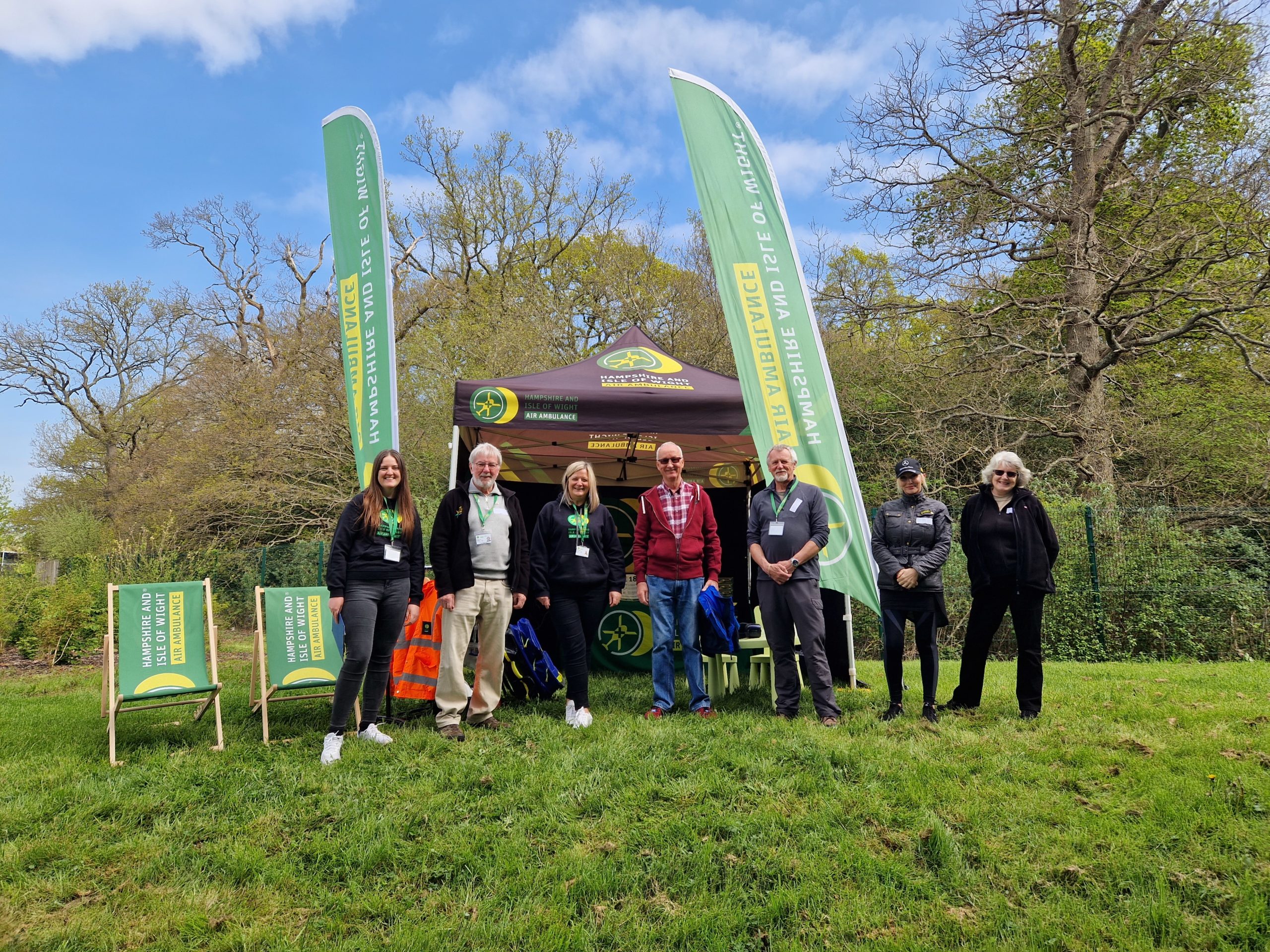 A group of people standing in a field in front of HIOWAA branded flags and gazebo.