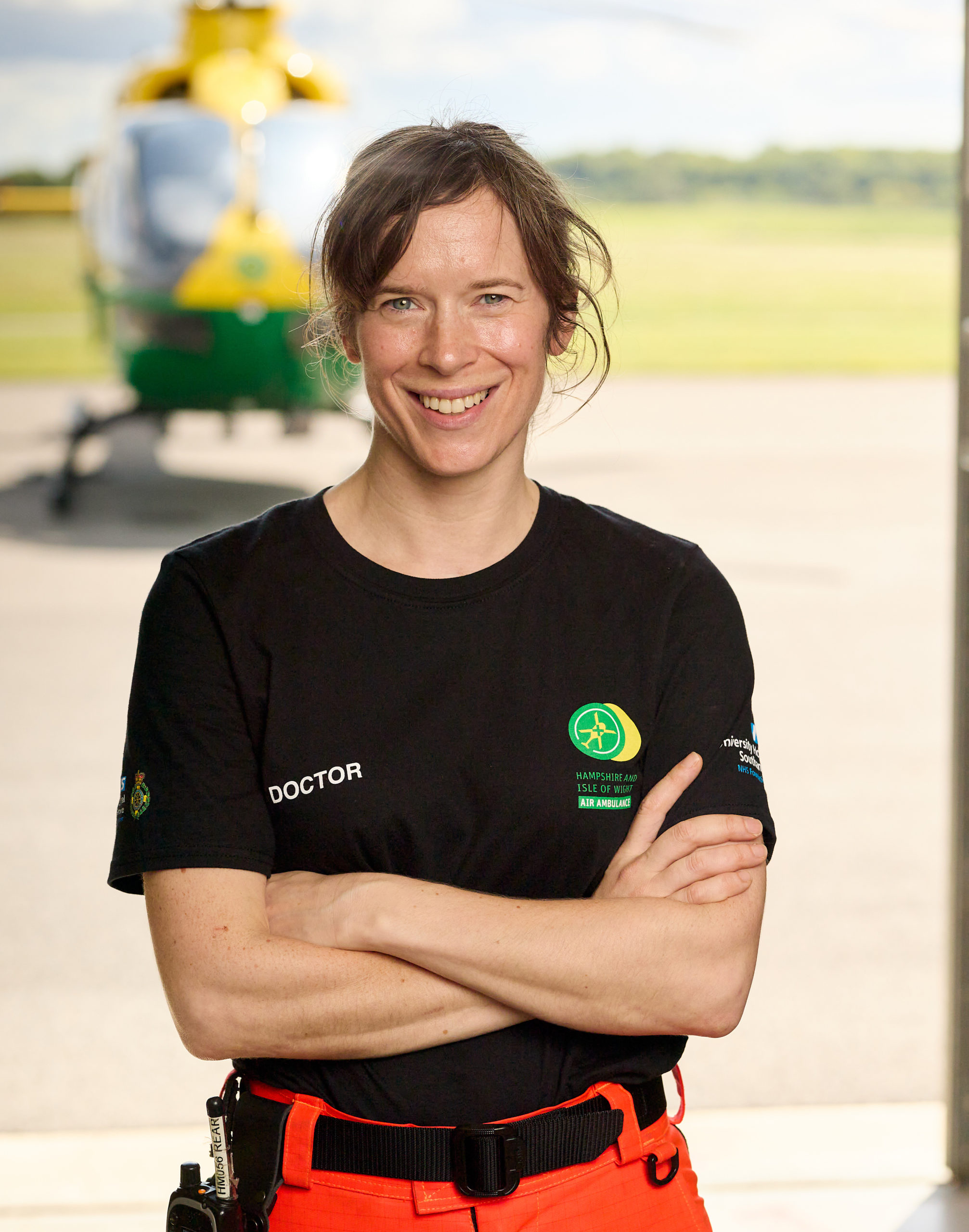 Dr Jen Hine stands in the hangar, smiling to camera. She has her arms crossed. The hangar doors are open and the helicopter is in the background.