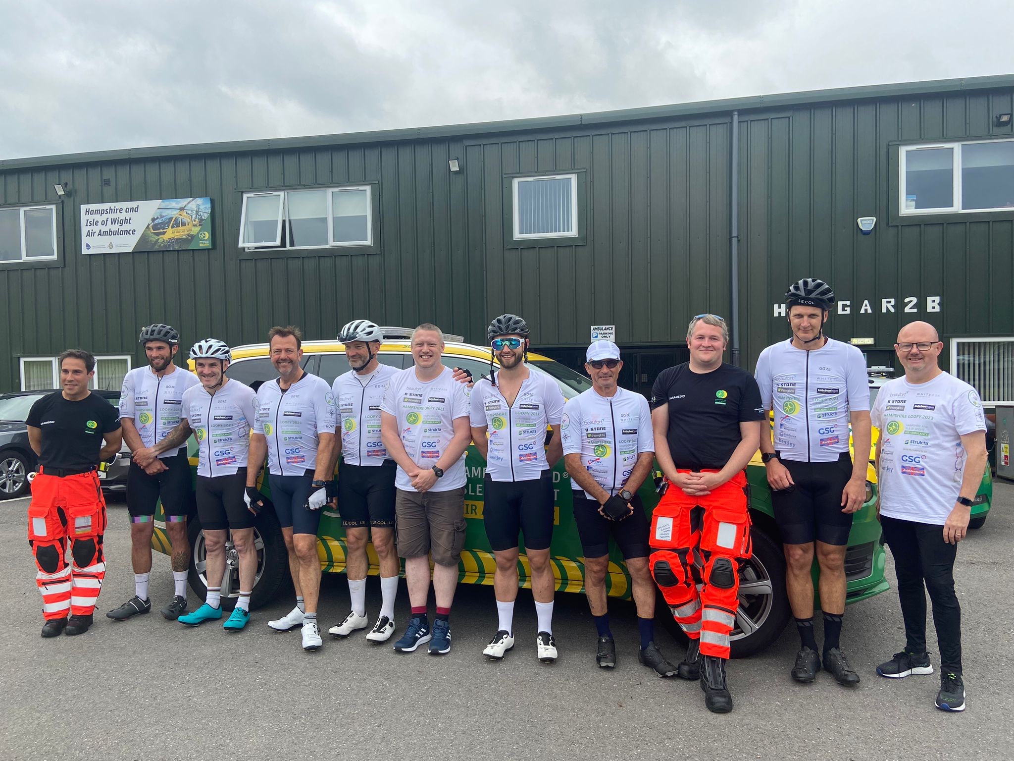 A group of cyclists and two paramedics standing in a line in front of an emergency response vehicle.