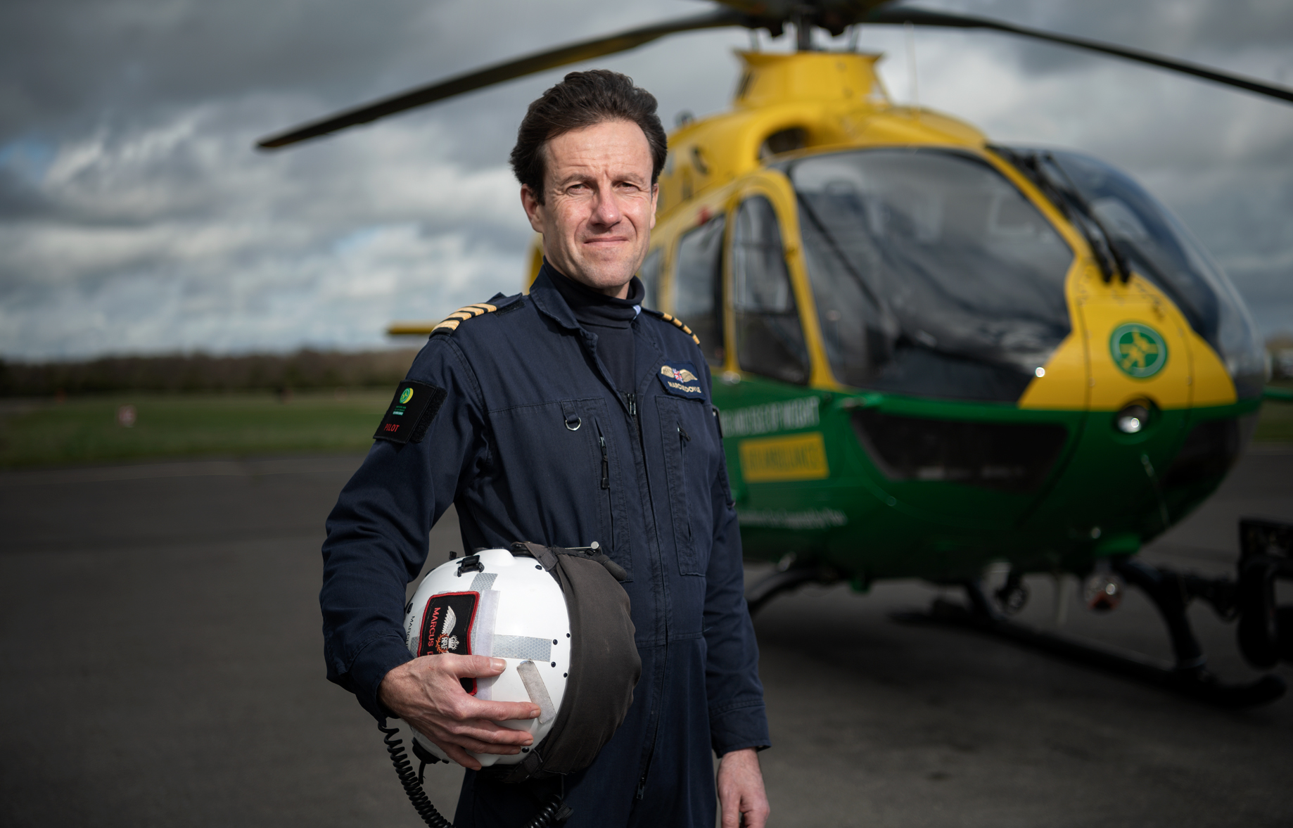 Pilot Marcus Doyle is standing in front of the helicopter. He is wearing his blue flight suit and carrying his flight helmet in his hands.