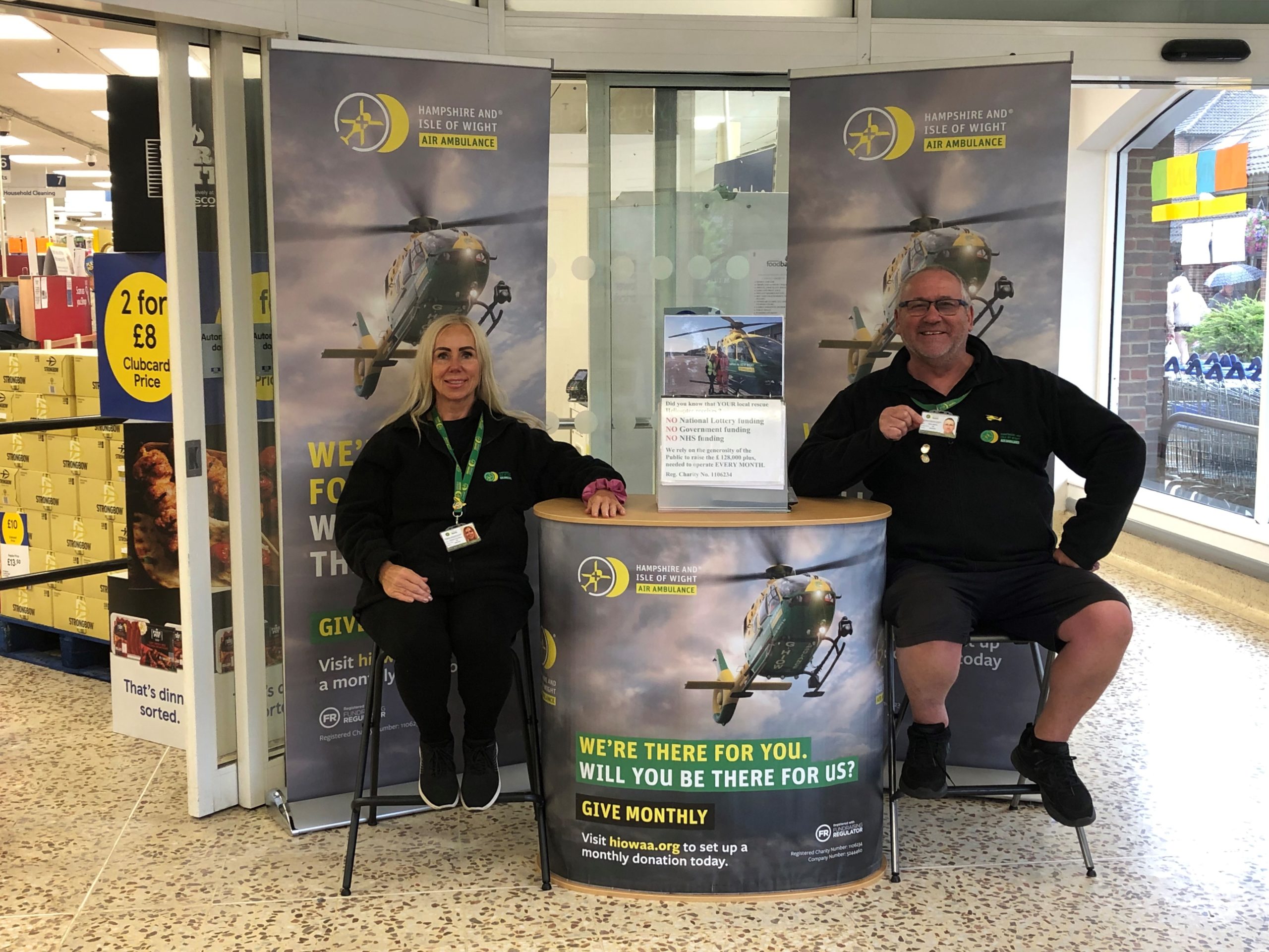 Mark and Theresa are sat in the foyer of a supermarket. They are surrounded by Hampshire and Isle of Wight Air Ambulance banners, and are leaning on a regular giving podium.
