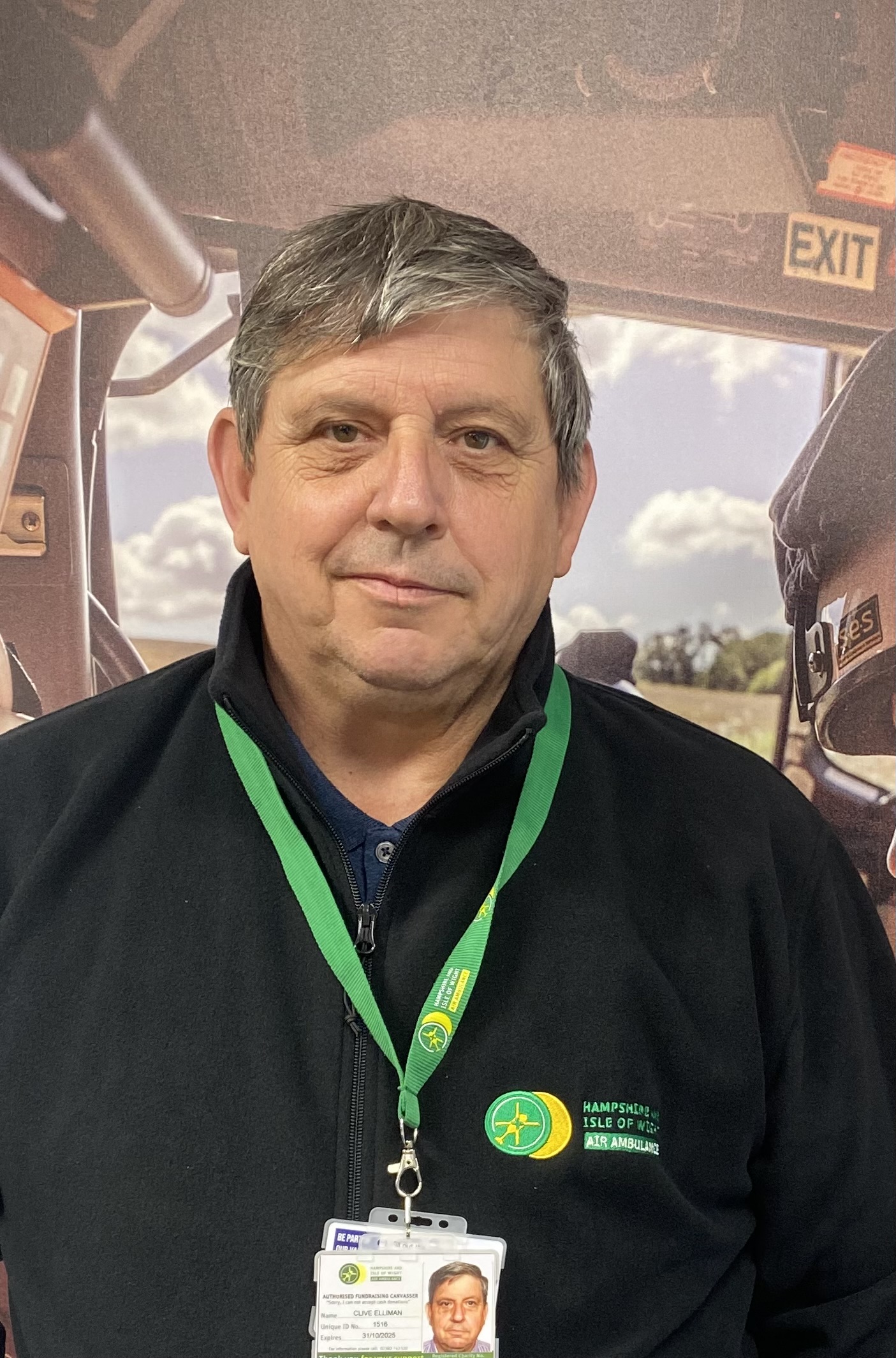 Clive is wearing a HIOWAA fleece and lanyard and standing in front of a large canvas photo of the crew