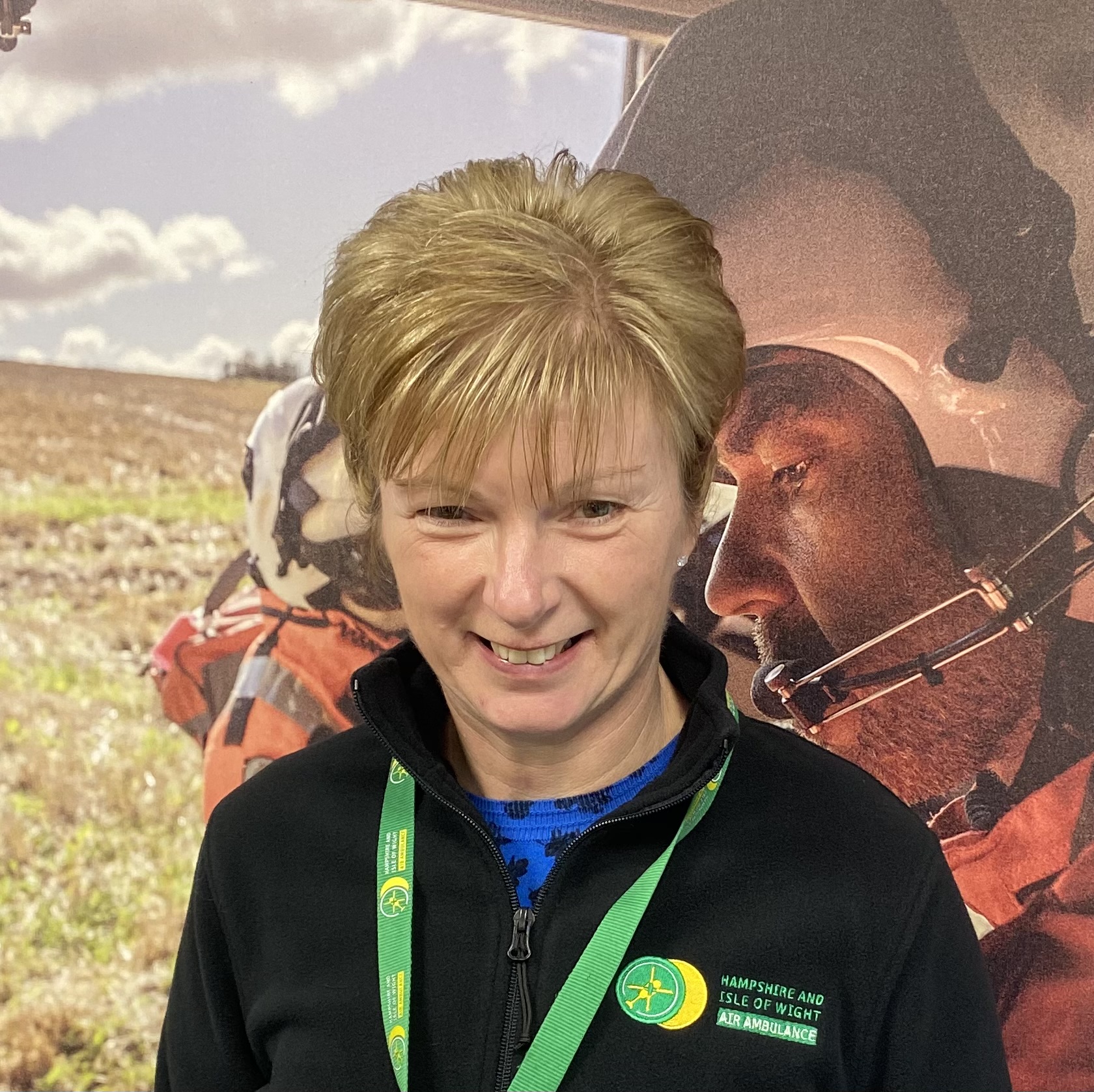Lisa is wearing a HIOWAA fleece and lanyard and standing in front of a large canvas photo of the crew