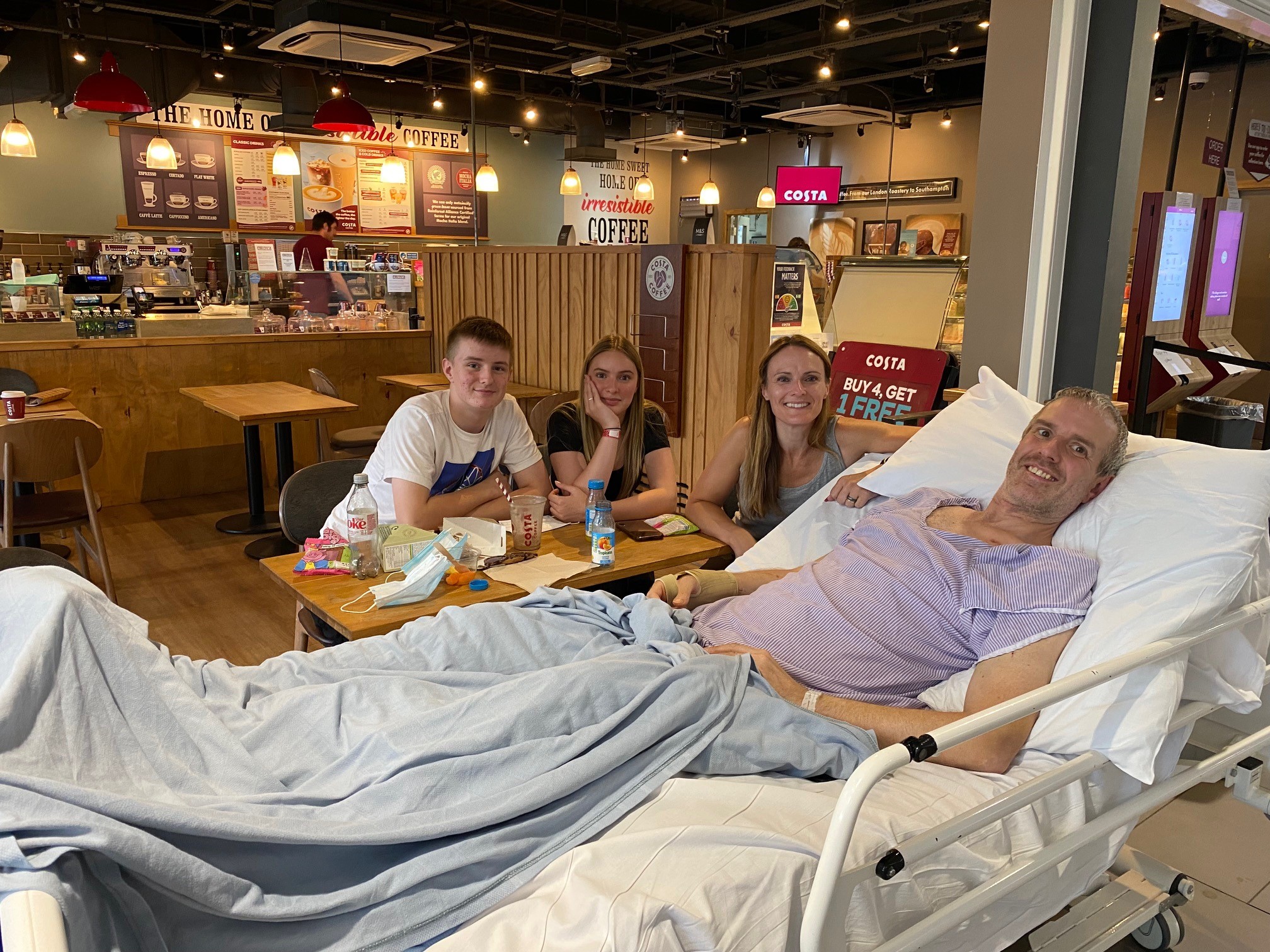 A family in a hospital coffee shop. The husband and father is lying in a hospital bed next to the table.