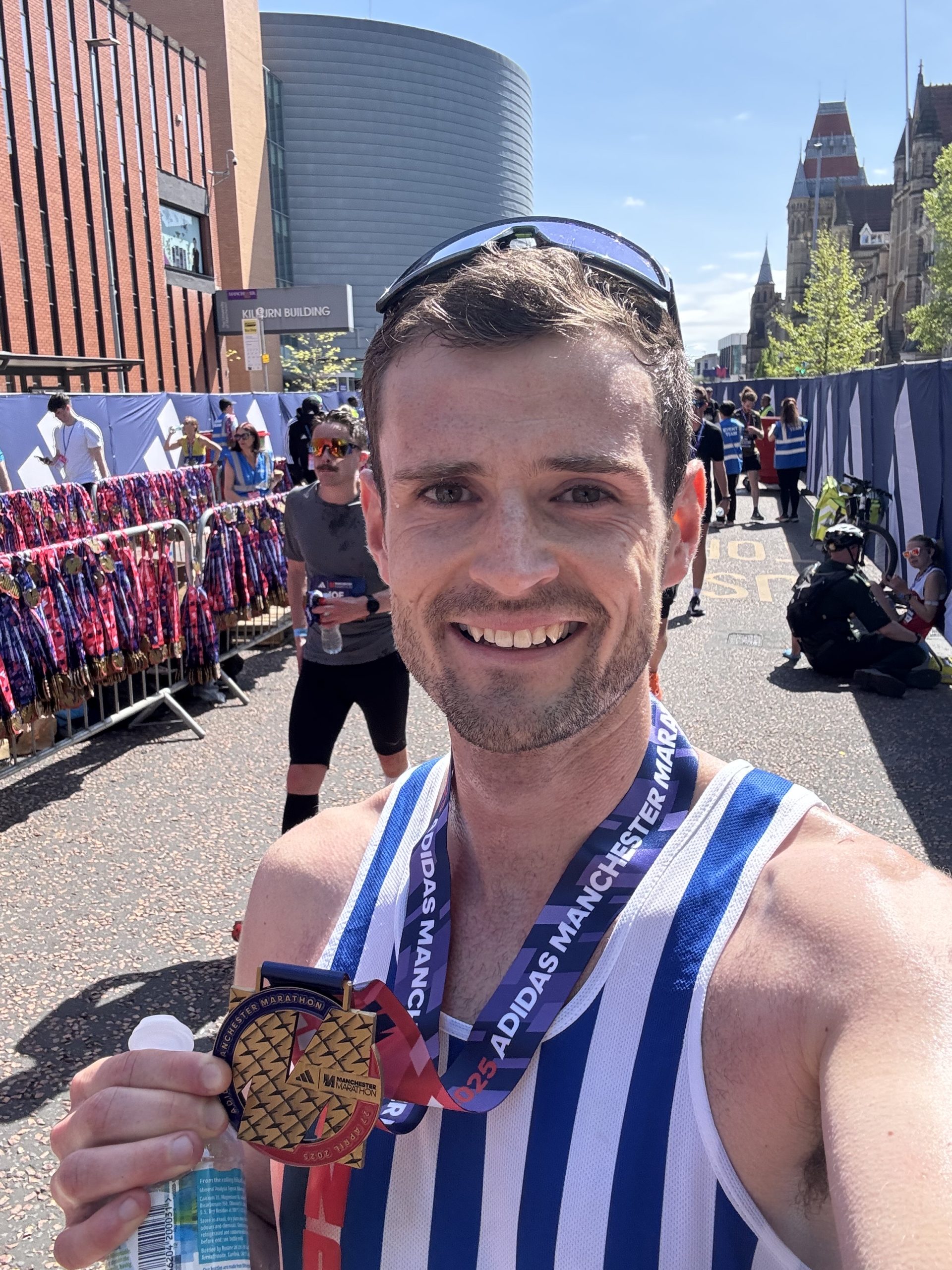 A man holding his Manchester Marathon finisher's medal. He is wearing a blue and white striped running vest.