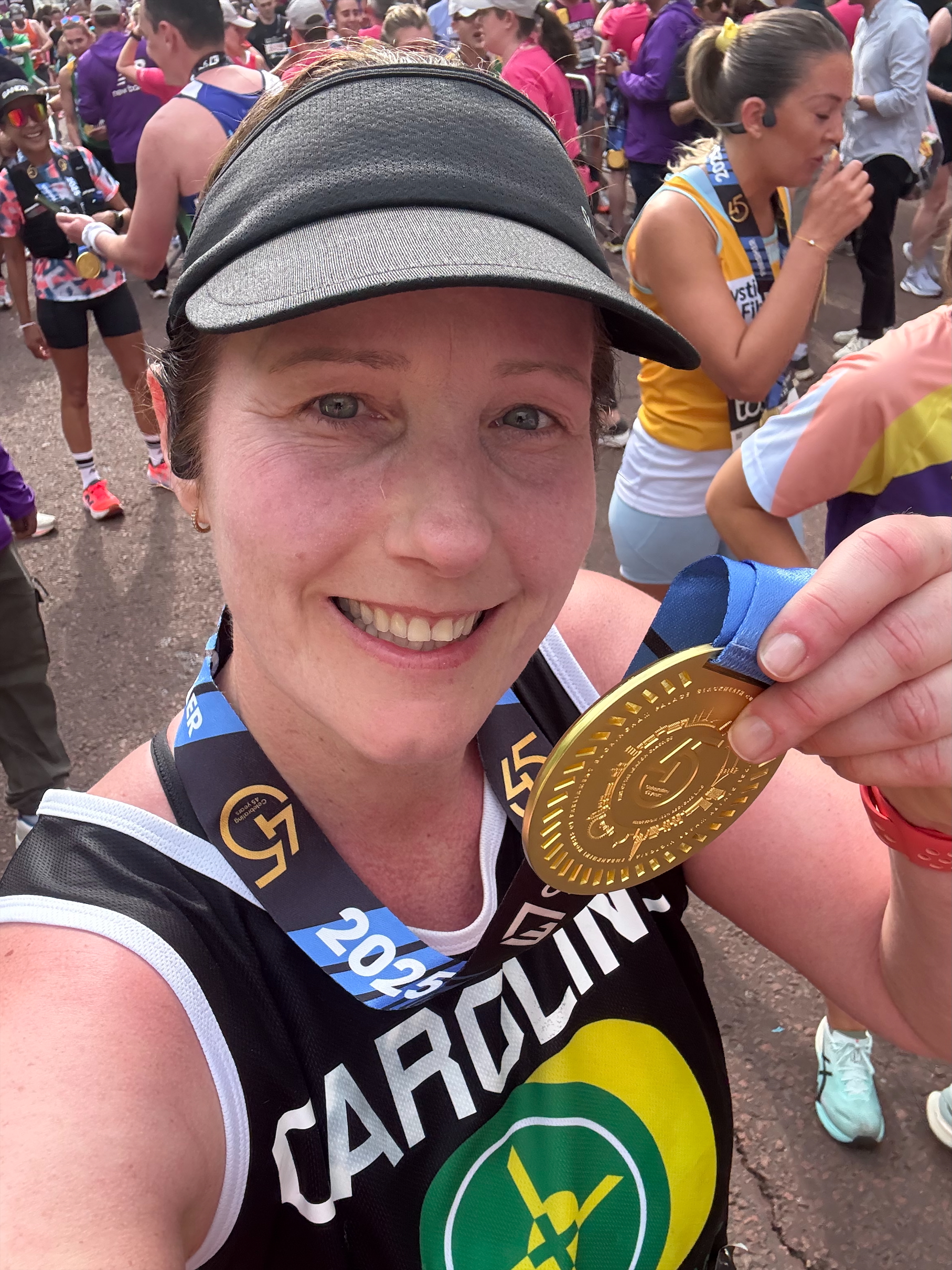 A lady holding up a London Marathon finisher's medal. She is wearing a HIOWAA running top which is black, green and yellow - it has Caroline written across the front.