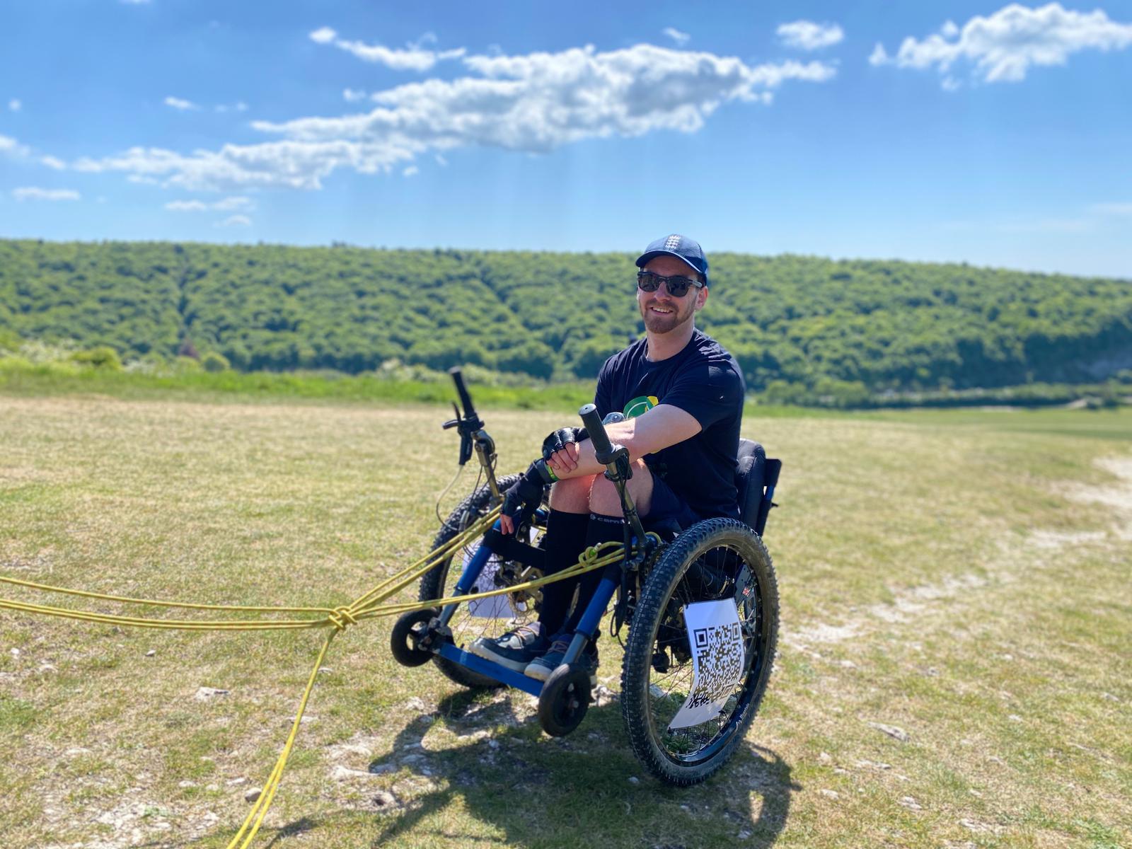 Dale on a practice climb. The sun is shining and he is up a hill. He has yellow ropes in front of him to help people pull him up in his wheelchair.