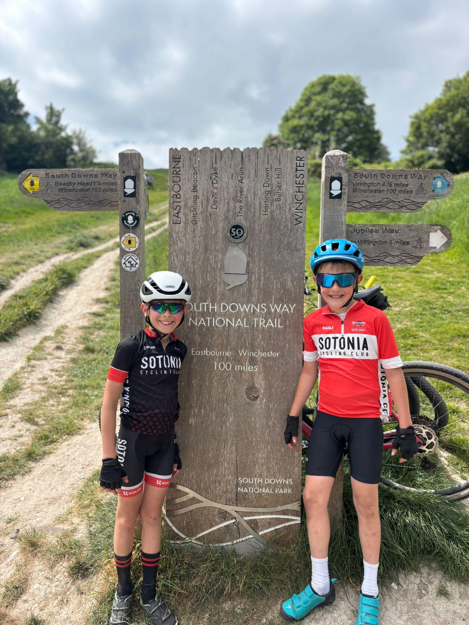 Two boys wearing cycling gear. They are stood next to a large wooden sign on the South Downs Way.
