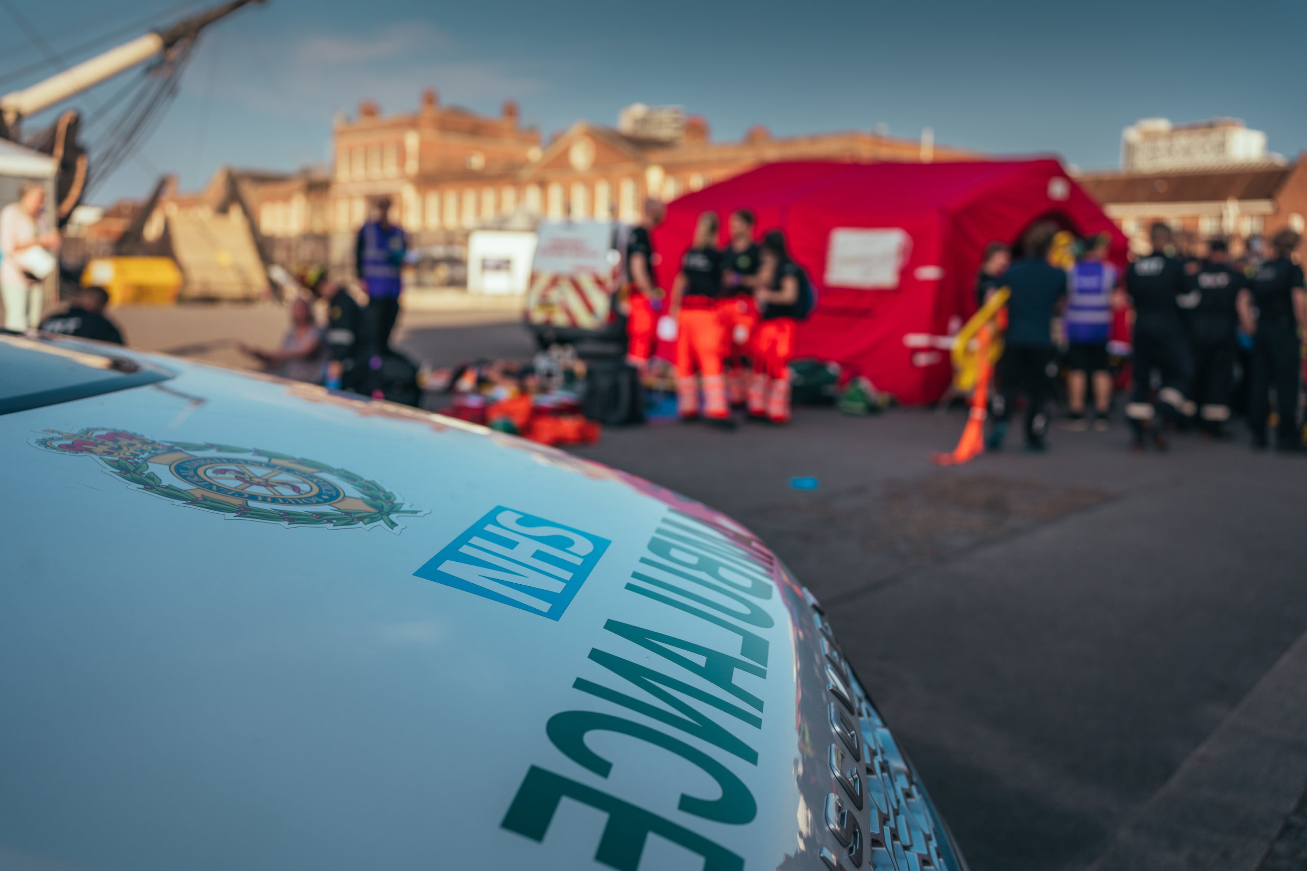 An NHS ambulance in the foreground. In the background are groups of emergency service staff.