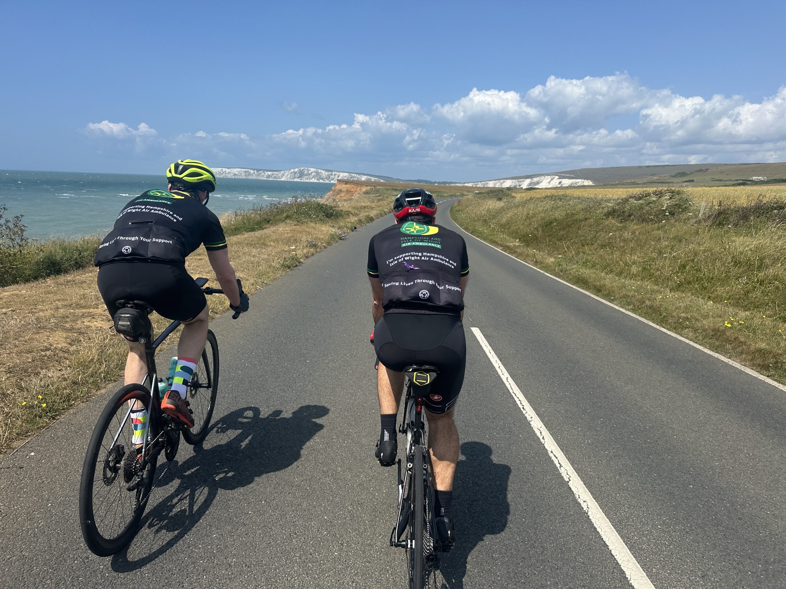 Two cyclists on a long road with cliffs and the coastline up ahead.