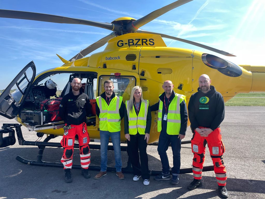 A group of people standing in front of a helicopter. Two of them are air ambulance crew members and three of them are visitors.