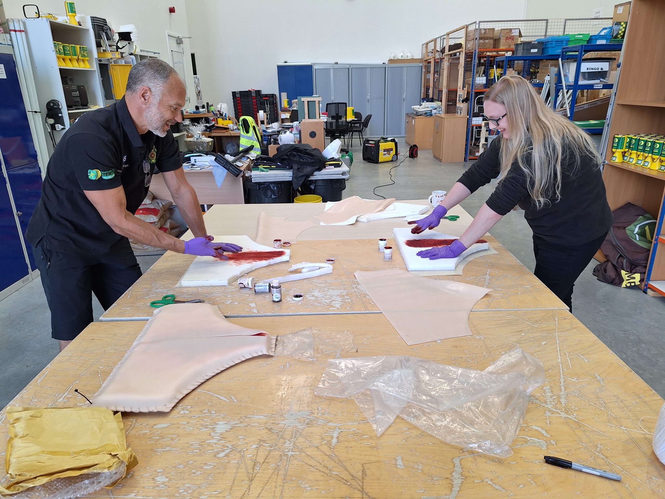 Two people are standing at a large table in a warehouse. They are making models for the purpose of emergency cesarean training.