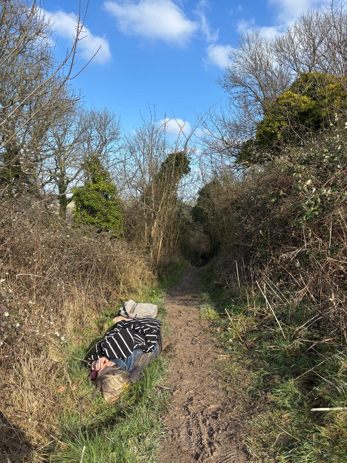 A lady lying on her side on a muddy path in a field. She is in pain and has her hands covering her face.