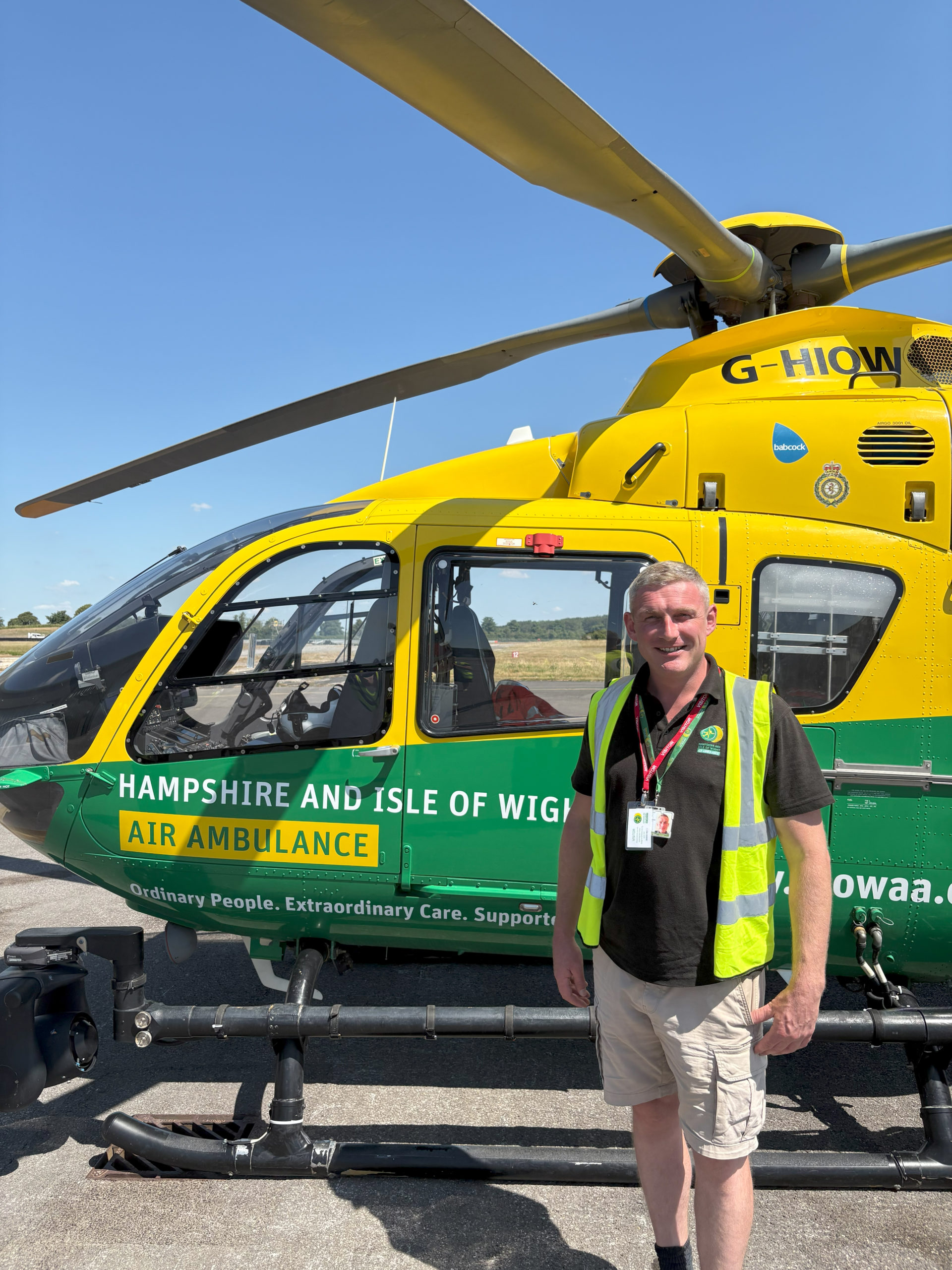 Mark is wearing a high visibility vest and standing in front of the green and yellow helicopter, smiling to the camera. It's a sunny day and the sky is blue,