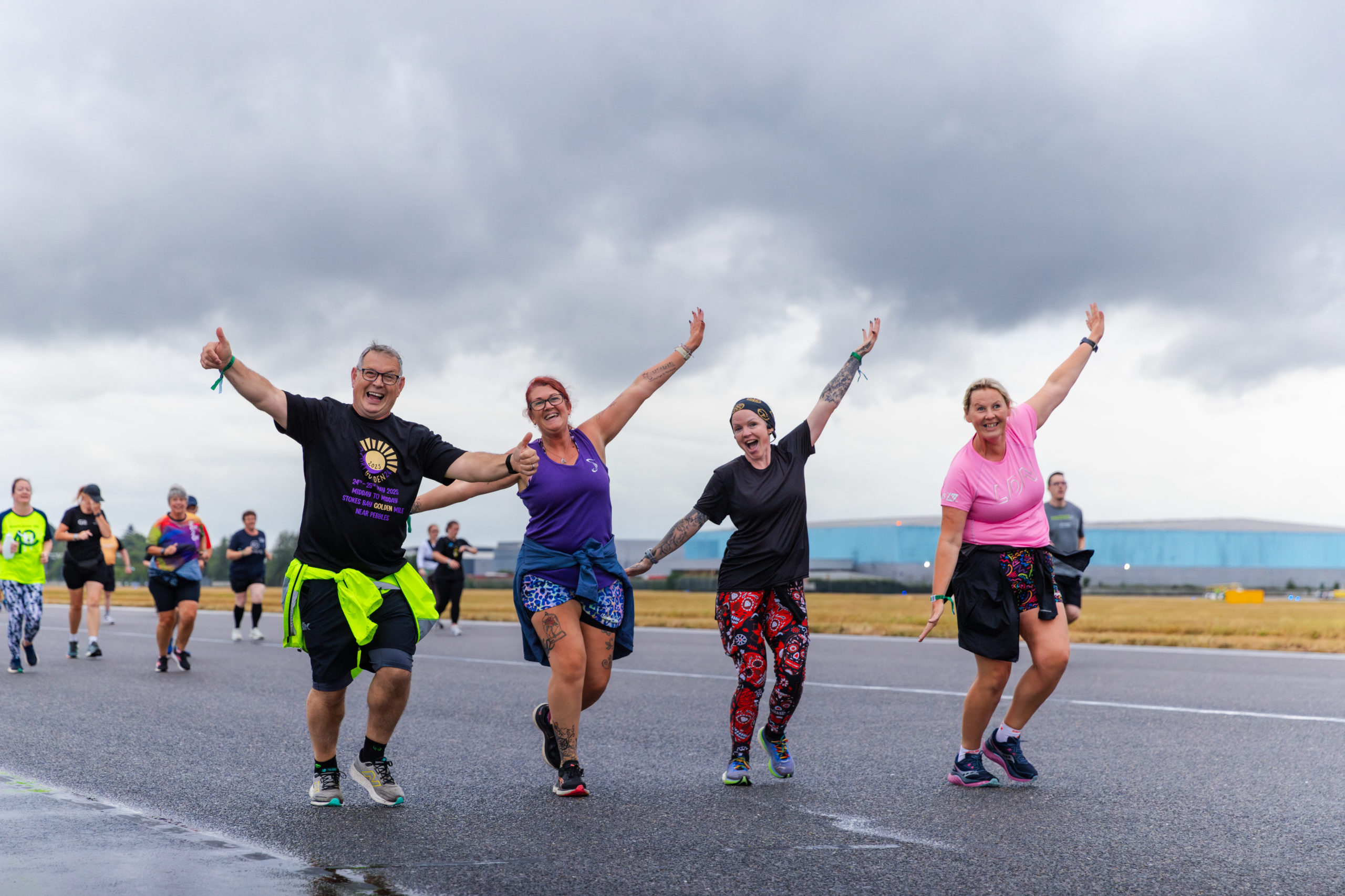 Four people running along an airport runway. They arms are outstretched as if they're flying like an aeroplane.