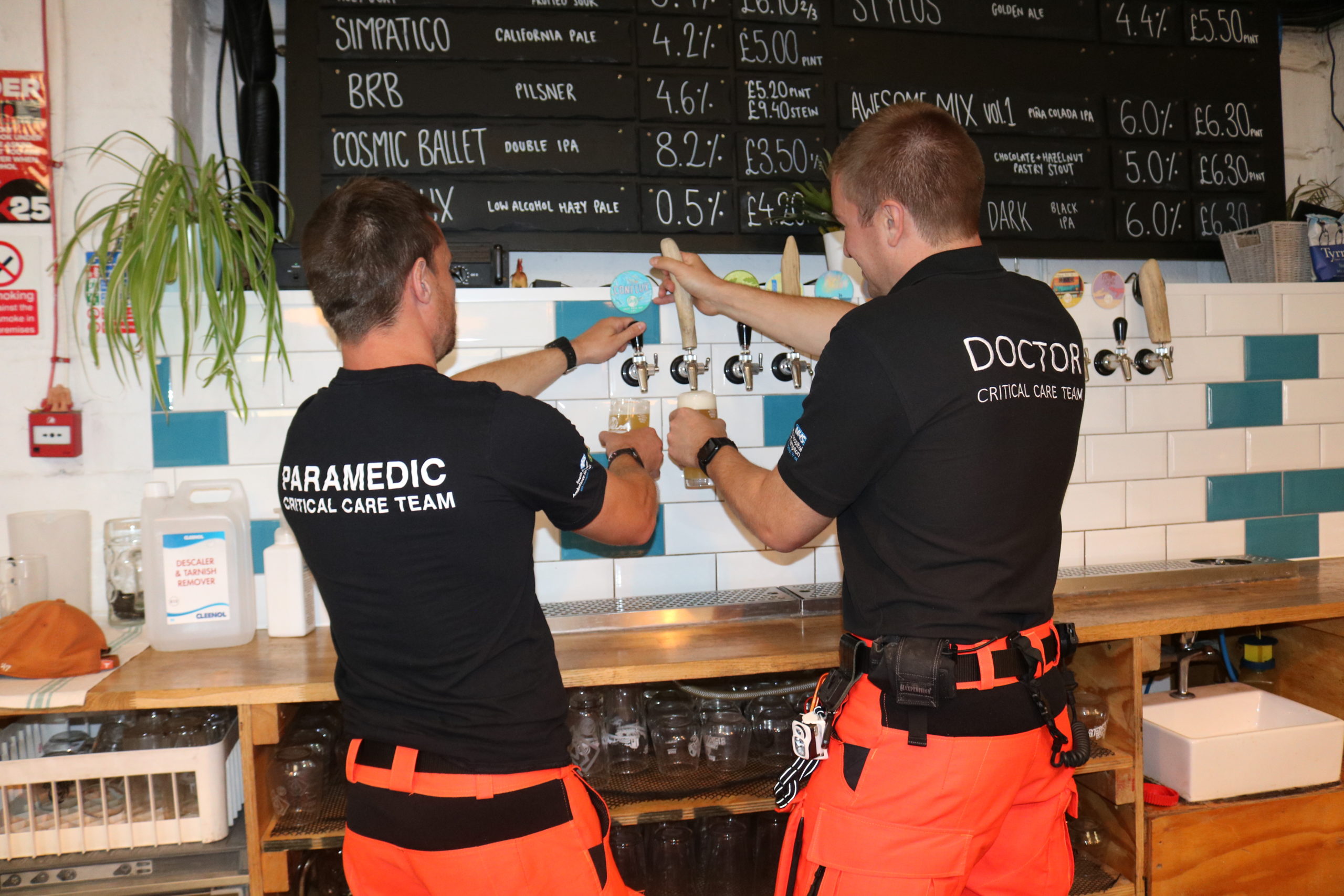 An air ambulance doctor and a paramedic pulling pints. Their backs are to the camera. They are wearing black T-shirts and orange flight trousers.