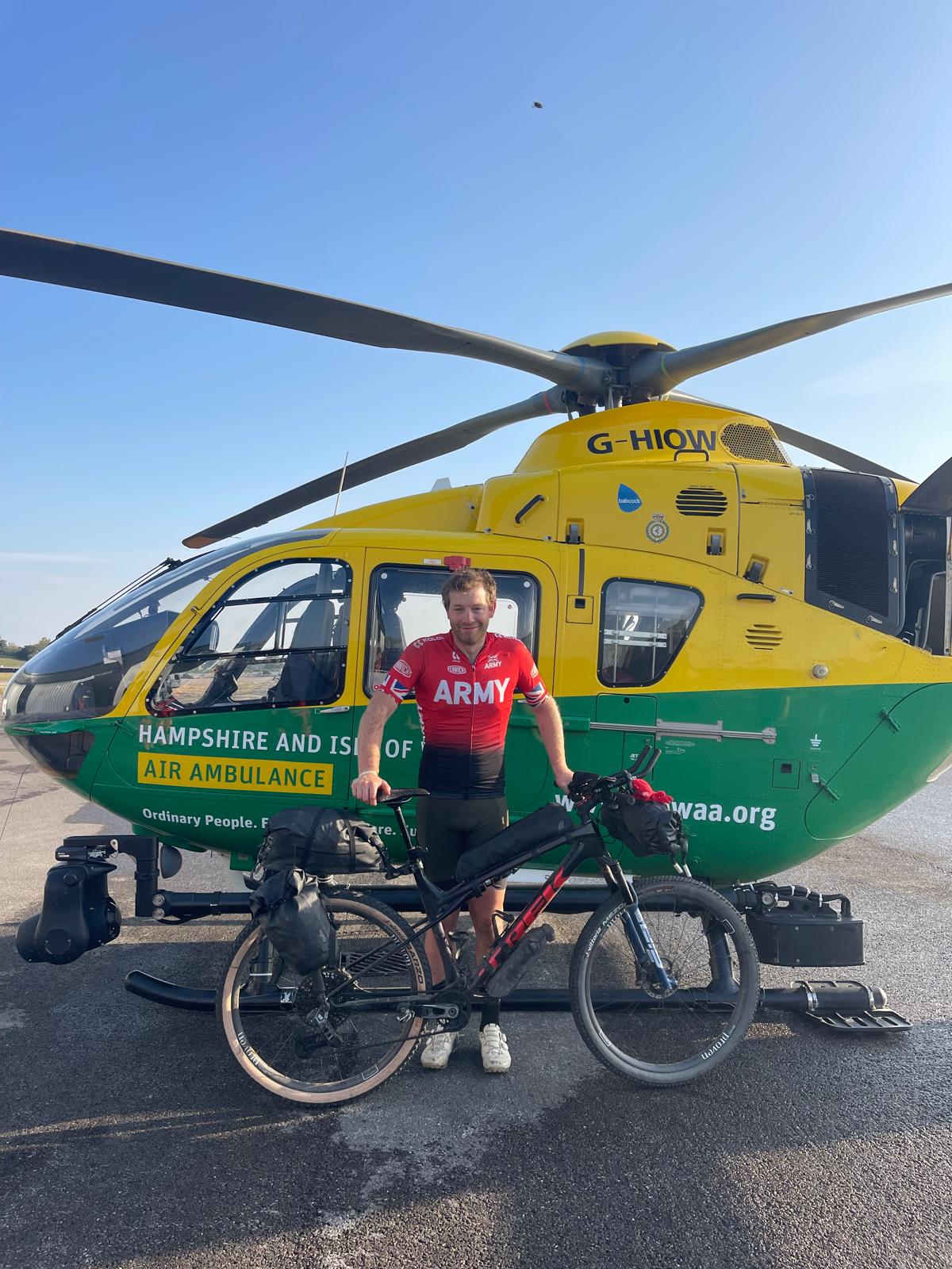 A man standing next to his road bicycle. He is standing in front of a green and yellow air ambulance helicopter.