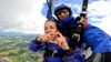 Two people tandem skydiving. The person strapped to the front is doing a love heart sign with her fingers.