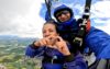 Two people tandem skydiving. The person strapped to the front is doing a love heart sign with her fingers.