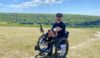 Dale on a practice climb. The sun is shining and he is up a hill. He has yellow ropes in front of him to help people pull him up in his wheelchair.