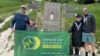 Four people stood by a large wooden sign that says "South Downs Way National Trail". They are holding a rectangular green banner with the Hampshire and Isle of Wight Air Ambulance logo.