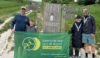 Four people stood by a large wooden sign that says "South Downs Way National Trail". They are holding a rectangular green banner with the Hampshire and Isle of Wight Air Ambulance logo.