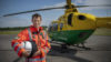 Dr Jonny Morris is standing in front of the helicopter wearing his orange flight suit. He is holding his flight helmet under his right arm and smiling to camera.