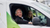 A man sat in a white and green van. He is in the driver's seat. His window is rolled down and his arm is resting on the door. There is a faint image of a woman sat in the passenger's seat.