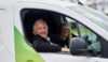 A man sat in a white and green van. He is in the driver's seat. His window is rolled down and his arm is resting on the door. There is a faint image of a woman sat in the passenger's seat.
