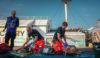 Two air ambulance crew members treating patients. Both patients are on stretchers on the ground, and the medical crew are knelt beside them. They are in a large outdoors area.