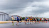 A group of people running on an airport runway. They are wearing running vests and T-shirts. There are dark clouds overhead.