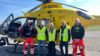 A group of people standing in front of a helicopter. Two of them are air ambulance crew members and three of them are visitors.